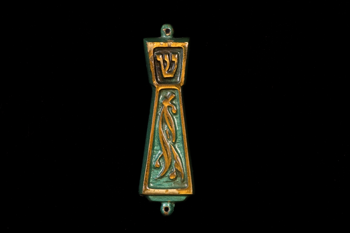 Mezuza tillverkad av metall med grön glasyr. Gyllene ornamentik. Innehåller pergamentrulle.