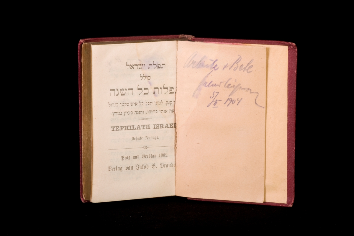 Siddur/bönbok skriven på papper med textilpärm och tillhörande pappfodral i vilket man kan skjuta in boken. Boken är skriven på hebreiska. Namn och årtal finns skrivet för hand på försättsbladet, och läses som "Arb.ite + Bete ……. 5/11 1904"