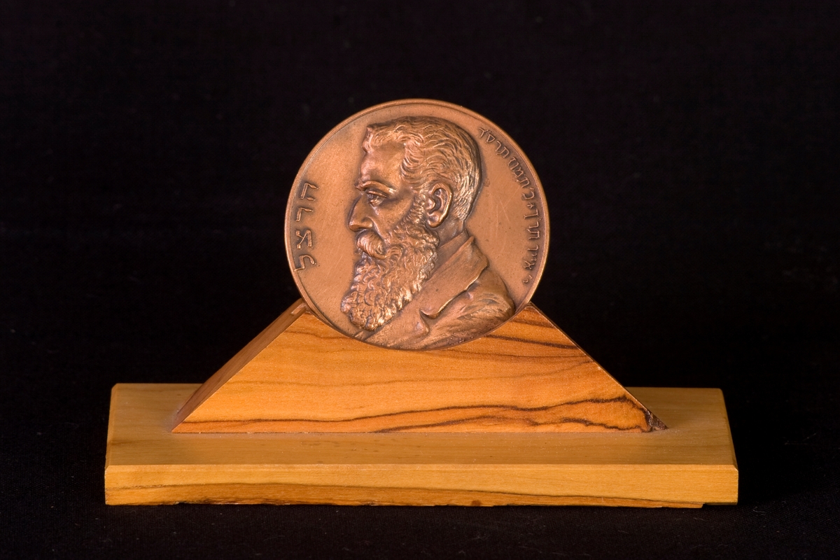 Medalj tillverkad av brons med ett tillhörande ställ av trä. Ena sidan dekorerad med engelsk och hebreisk text, och andra sidan dekorerad med Theodor Herzel i profil samt hebreisk text.
