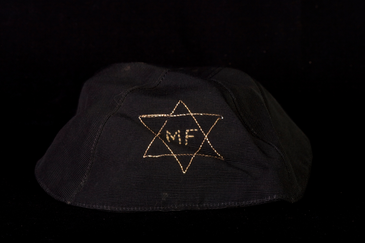 Kipa tillverkad av svart textil. Dekorerad med en davidsstjärna med initialerna MF inuti, broderade med guldfärgad tråd.