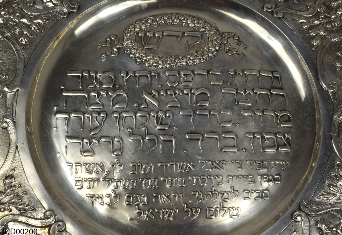 Stort silverfat tillverkat för pesach. Fatet är rikt dekorerat med präglad dekor i form av hebreisk text på spegeln samt blommor, frukt, personer (Aron, David, Mose) och olika scener på kanten. Vågig kant.