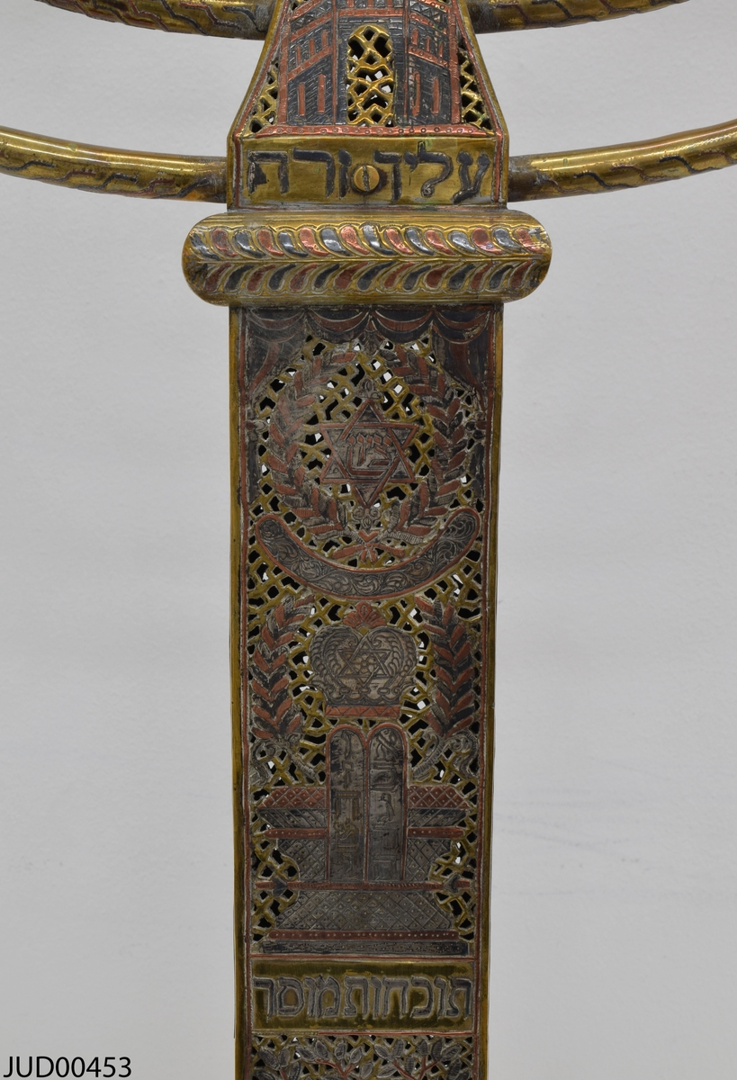 Stor chanukkaljusstake tillverkad av mässing, i form av en obelisk dekorerad genombruten dekor i rött och svart. Davidsstjärna på toppen mellan ljushållarna.