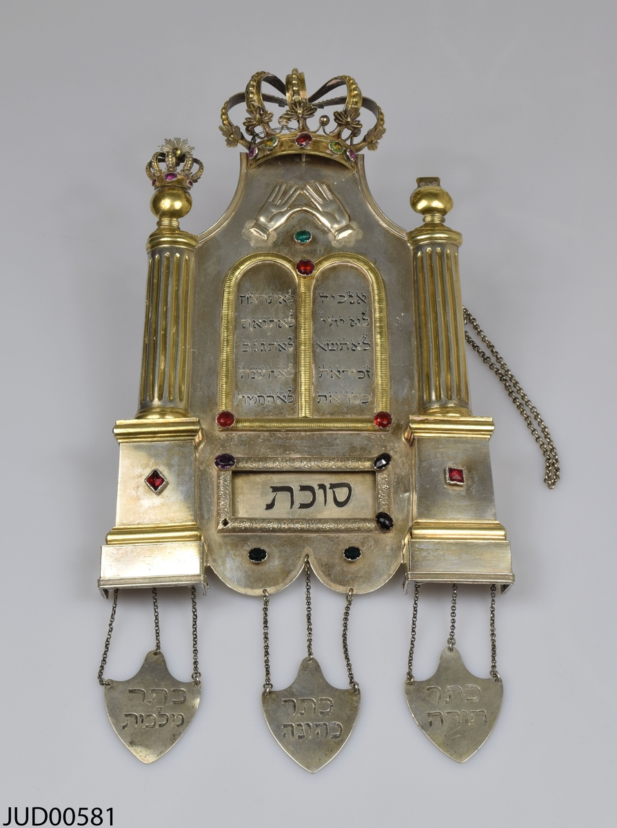 Torahsköld med två torahpekare liggandes i låda. Lådan är klädd med svart skinn på utsidan och beige sammet på insidan. 

Skölden är tillverkad av silver och dekorerad med färgade stenar. Kolonner på sidorna och krona på toppen, samt tre hängande plaketter i nederkant. 

Pekarna är tillverkade av silver. En är vriden och utan annan dekor, medan den andra har en rak fyrkantig form och dekorerad med ristad hebreisk text.