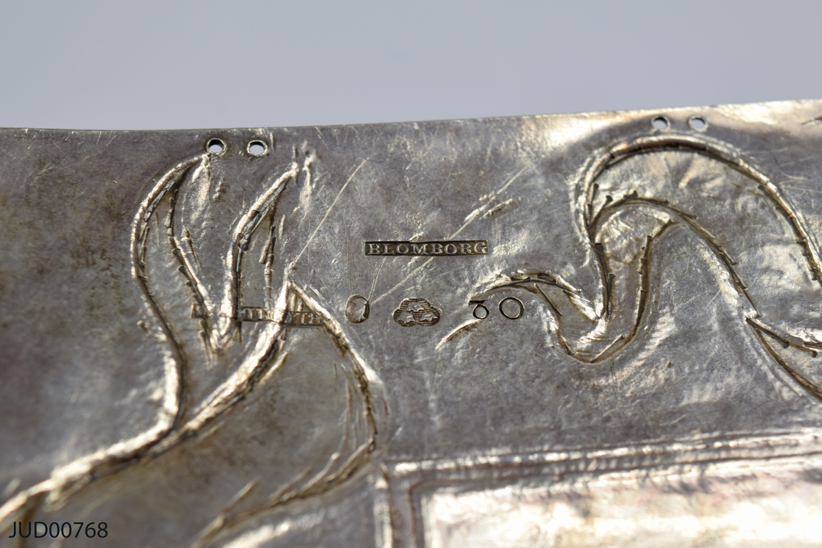 Torahsköld tillverkad av silver. Detaljer på sköldens framsida med lilla riksvapnet i mitten, och stiliserade stentavlor undertill med hebreisk text. Runt om girlanger med hebreiskt text inuti.