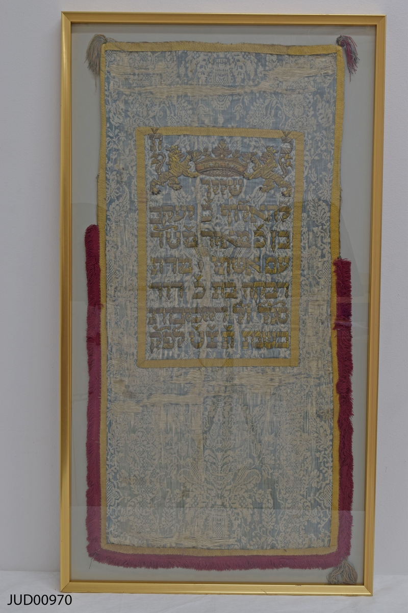 Torahförhänge i siden med röd frans nedtill och text i mitten.