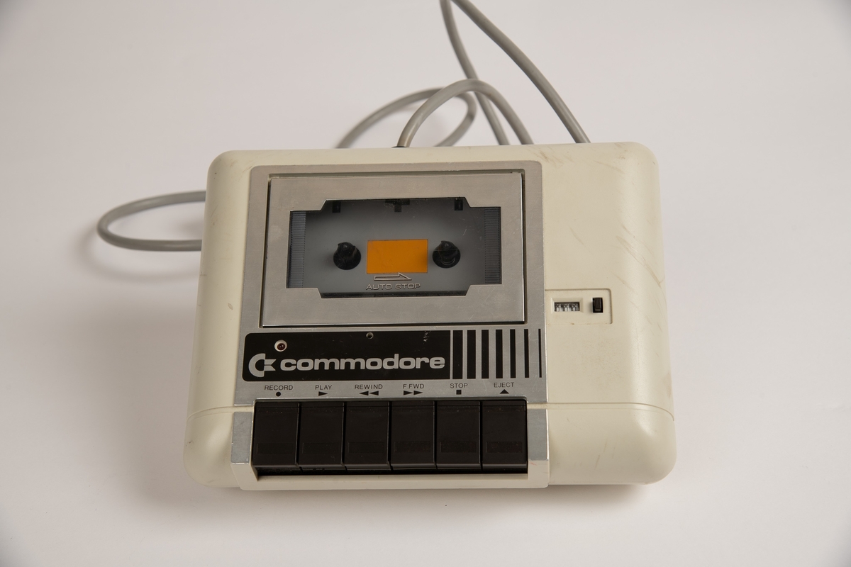 "Kassettspiller" til Commodore 64. Tilkoblet er en grå ledning som skal kobles i Commodore 64 tastaturet. 
Kanpper til "record", "play", "rewind", "fast forward (f. fwd)", "stop" og "eject".