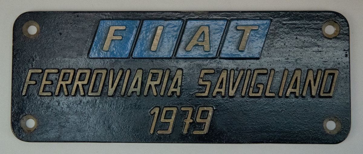Tillverkarskylt av aluminium med svart botten och texten "FIAT" i blå rombformiga fält högst upp och nedanför "FERROVIARIA SAVIGLIANO 1979". Skylten har formen av en liggande rektangel med rundade hört och har ett nedsänkt skruvhål i vardera hörn.