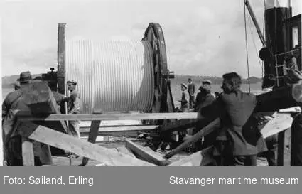 Kablenedlegging utenfor Hundvåg. Arbeider med å legge elektrisk kabel fra båten Rosenberg. Kablerull merket Skandinaviske fabrikker i Norge - Oslo. 
Flere tilskuere og arbeidsfolk i robåter