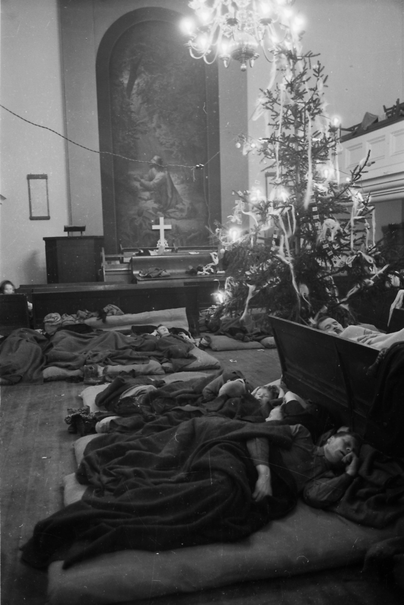 Personer sover i Metodistkirkenen i Trondheim i jula 1944. De ligger på madrasser på gulvet og på kirkebenker. De har enkle pledd å dekke seg til med. I bakgrunnen ser man prekestolen, alteret med altertavle, et juletre og galleriet.