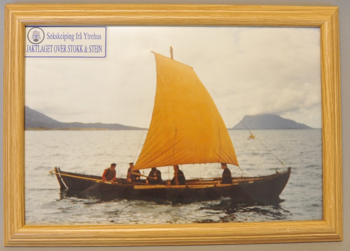 Innrammet foto av en båt med årer og seil. Fem personer ombord i båten. Fjord og fjell i bakgrunnen