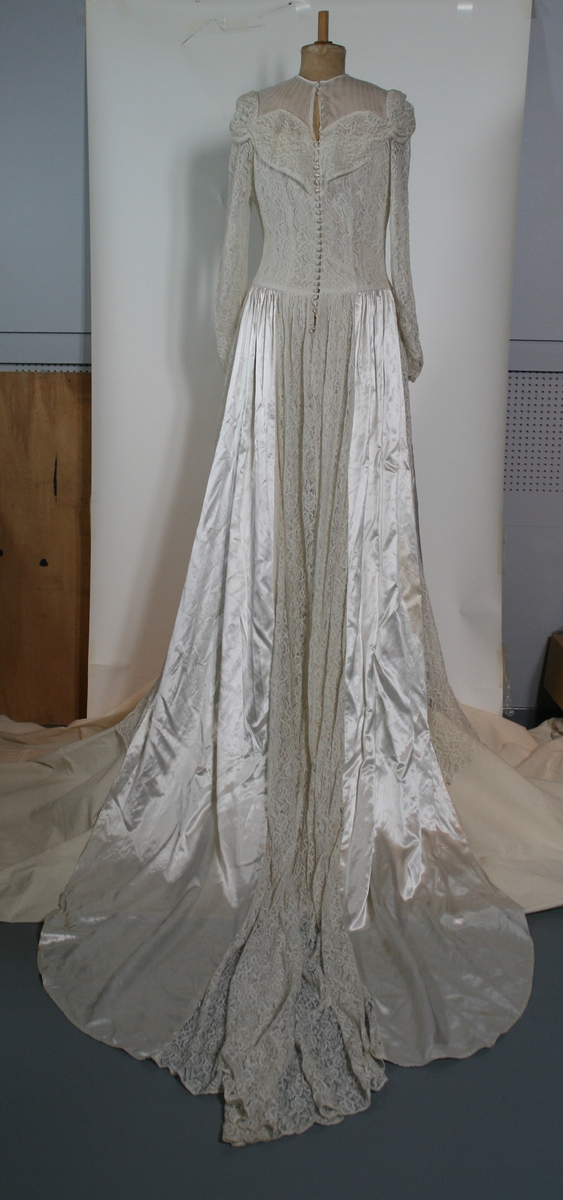 Brudekjole med lange ermer og slep. Overdelen og ermene er i formsydd med blonder over et silke-lignende tekstil. Skjørtet har blonder på front- og bak-parti, med silke på sidene. Knapper i ryggen.