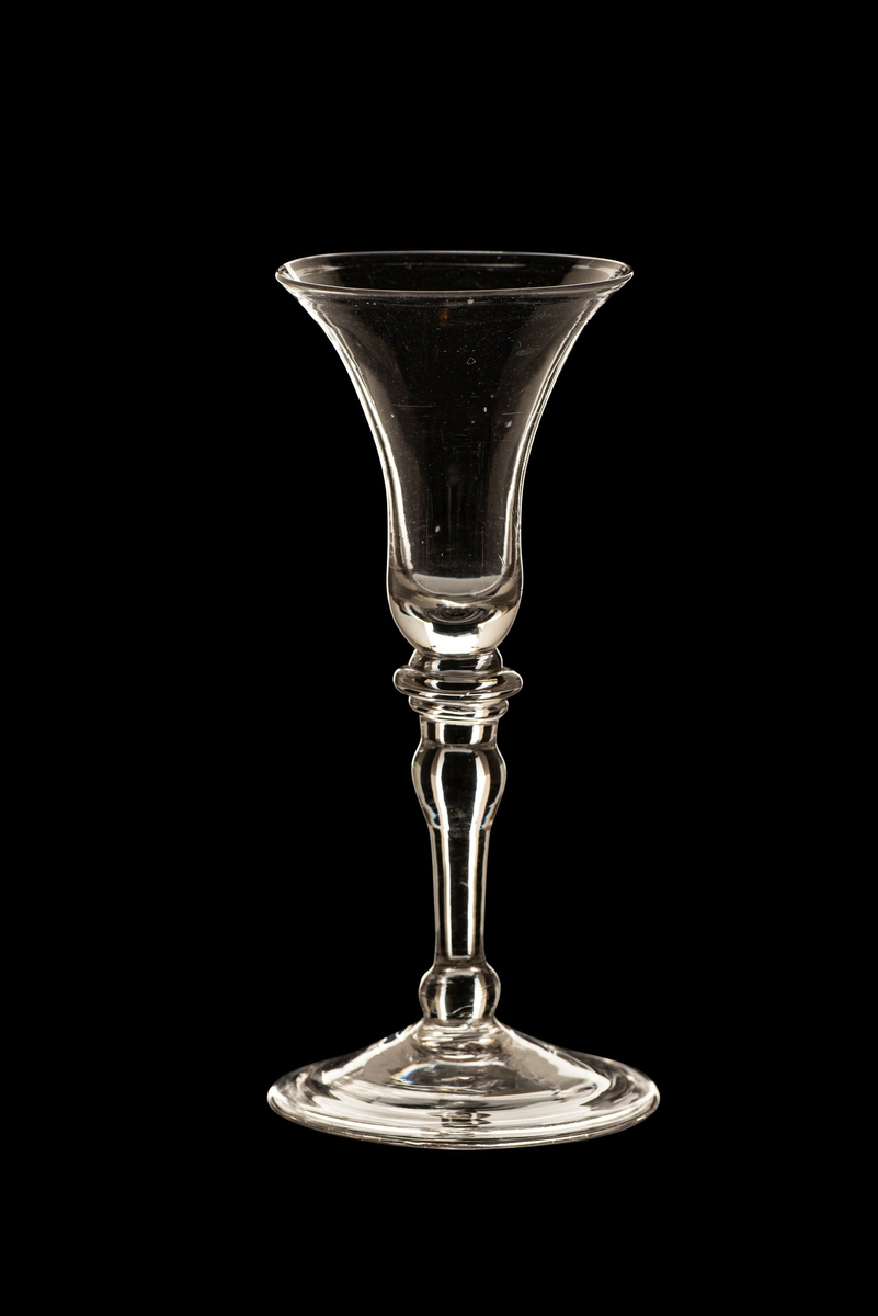 Brennevinsglass, variant av "Trundhiems Brendevins Spits Glas". Rent, tilnærmet fargeløst glass. Trompetformet klokke med slank, balusterformet stett. To skiver under klokken og kuler i stettens ender. Ombøyet fotkant. 