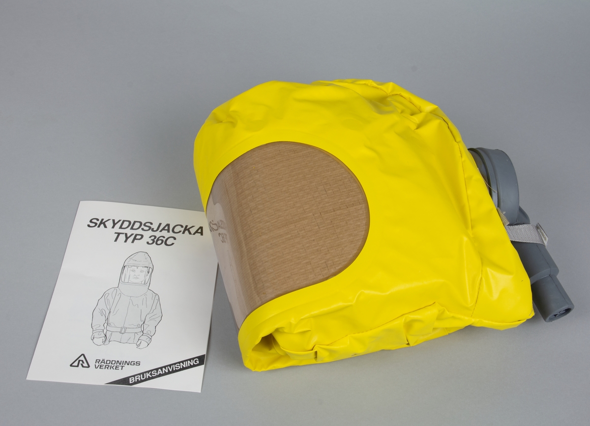 Kartong med lock och handtag innehållande skyddsjacka Typ 36C. I kartongen finns en oöppnad förpackning i plast med en gul skyddsjacka, jackan har ett omslag av oblekt silkespapper. I påsen ligger en bruksanvisning från Räddningsverket.
