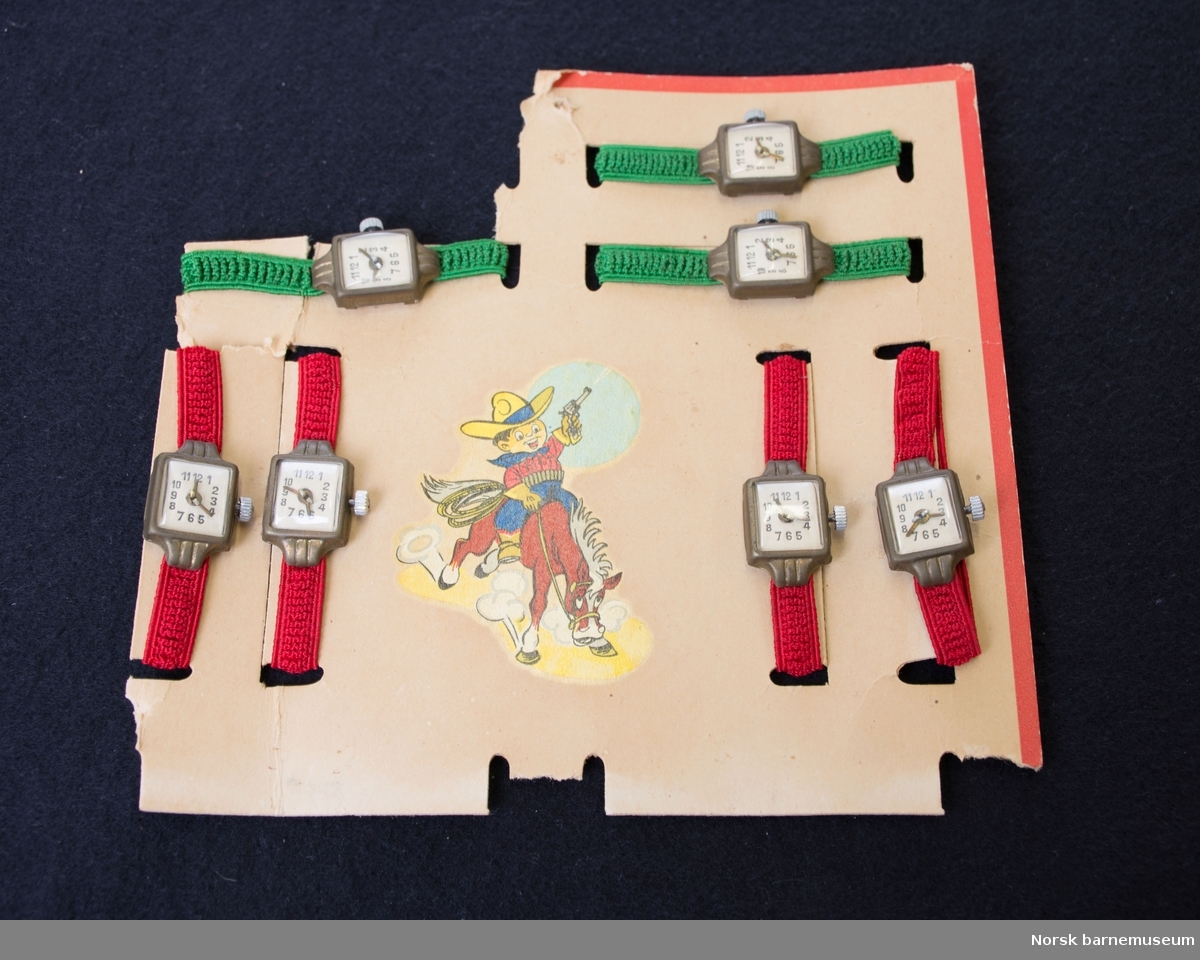 7 lekeklokker med strikk til å ha rundt hånden er samlet på et brett. 3 av klokkene har grønn strikk, og 4 har rød strikk. 