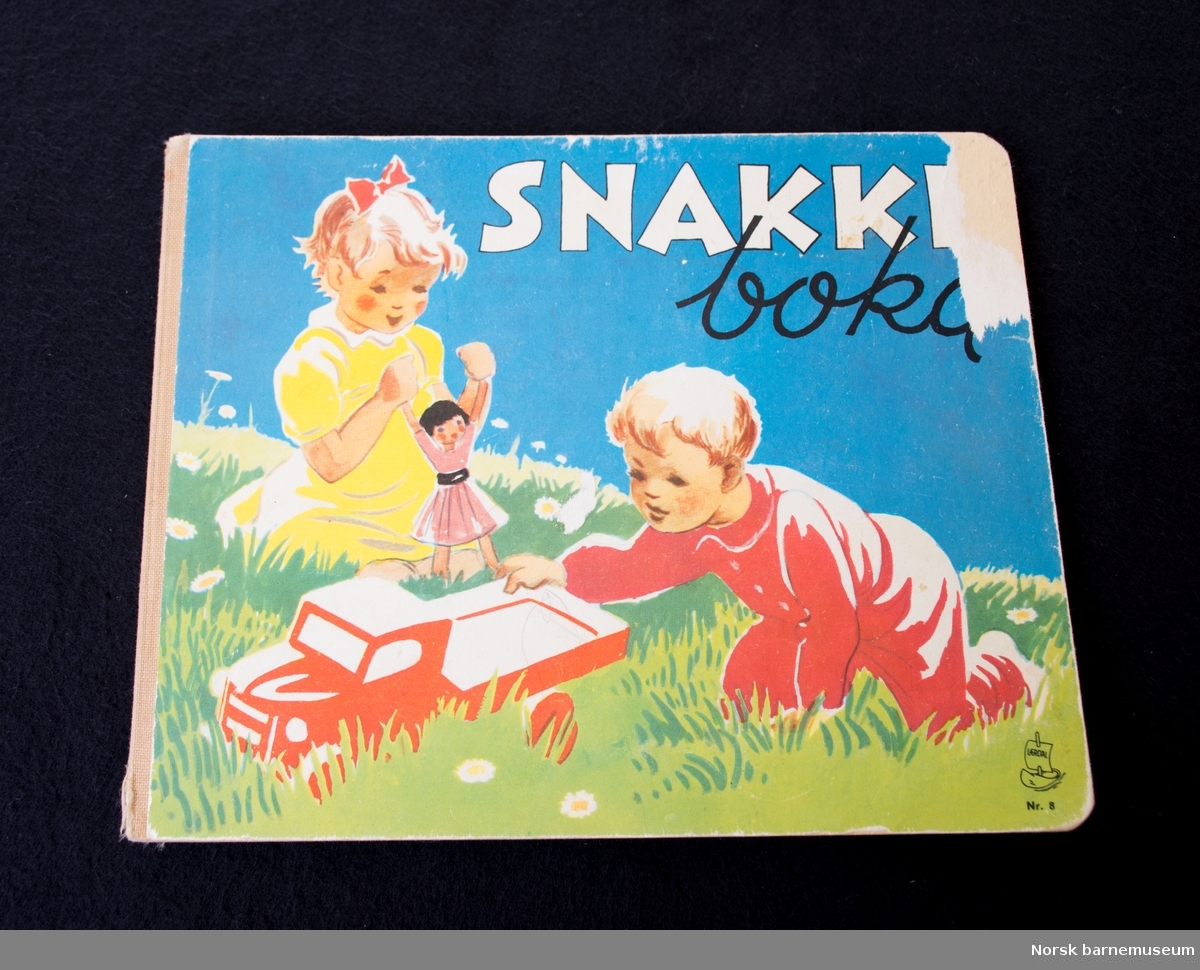 Fremsiden av boken har et bilde av to barn som leker. Jenta leker med en dukke og gutten leker med en bil. 
