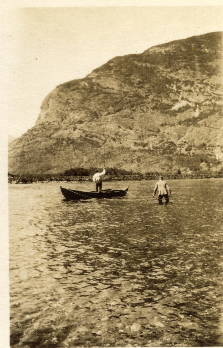 To menn fisker i Lærdalselvi, Sogn og Fjordane. Den ene fisker med stang fra en robåt, den andre står i elven og kontrollerer båten med tau. Fotografert 1916.