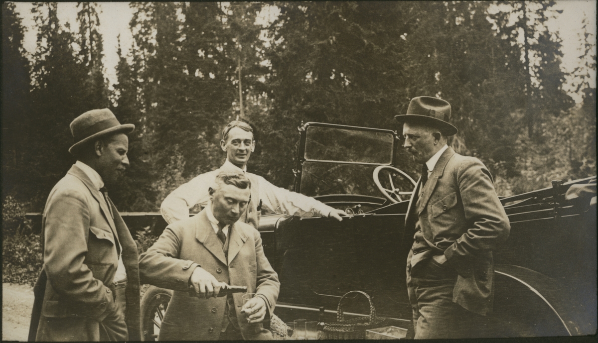 Fire menn ved åpen bil, i forbindelse inspeksjon av tømmerfløting. Carl "Ullern" Løvenskiold åpner en flaske. Fotografert 1922.