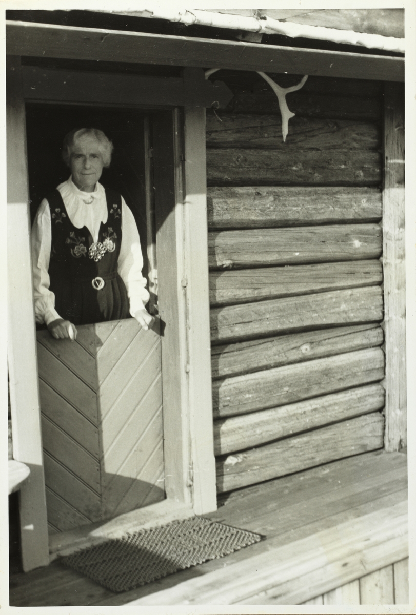 Olivia Hansen, ikledd bunad, i døren til tømmerhytte. Fotografert 1953.