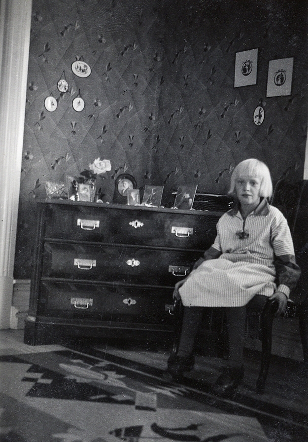 En flicka sitter på en stol bredvid en byrå. På byrån syns några foton. 
Under fotot text: "Hos Gerda 1931".