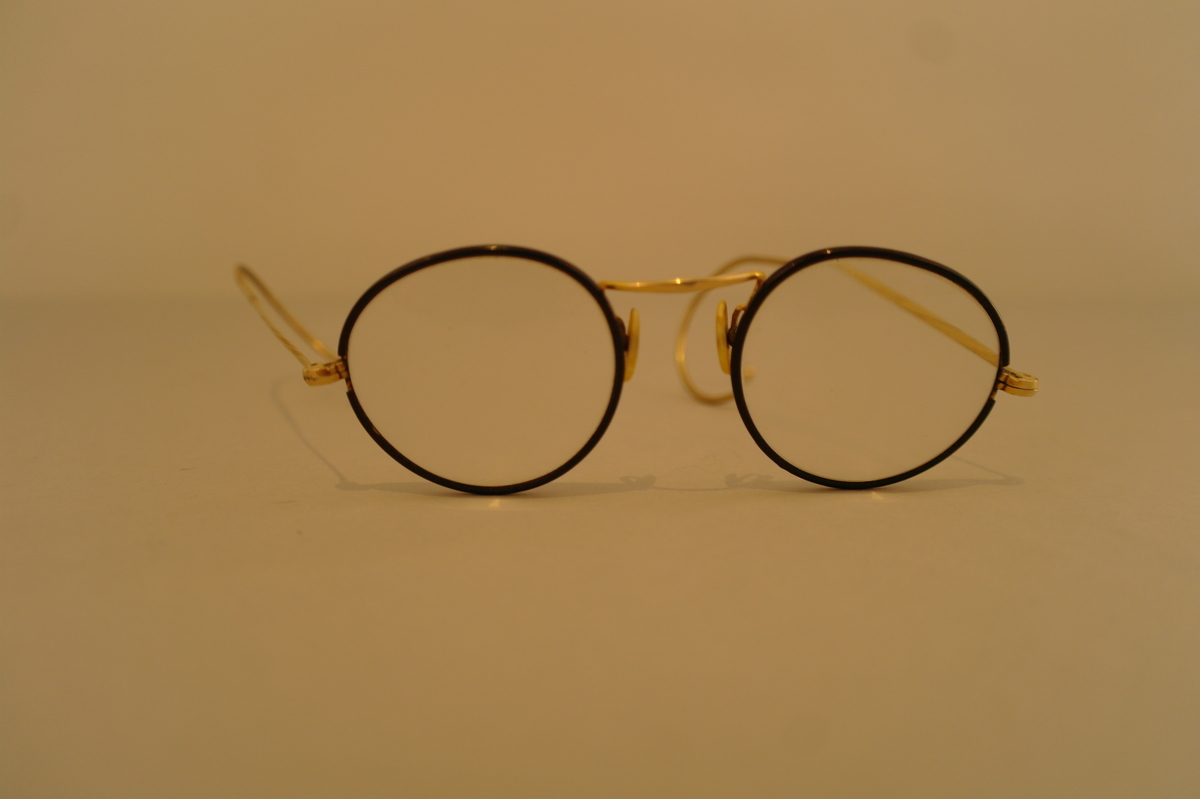 Brillehus med briller. Brilleglasene er rund og har ein svart infatning og med stenger som kan festes bak øyrane.