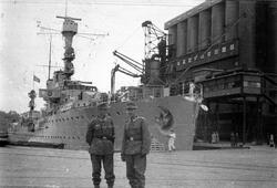 To soldater står på en kai foran et krigsskip. Kan se ut som