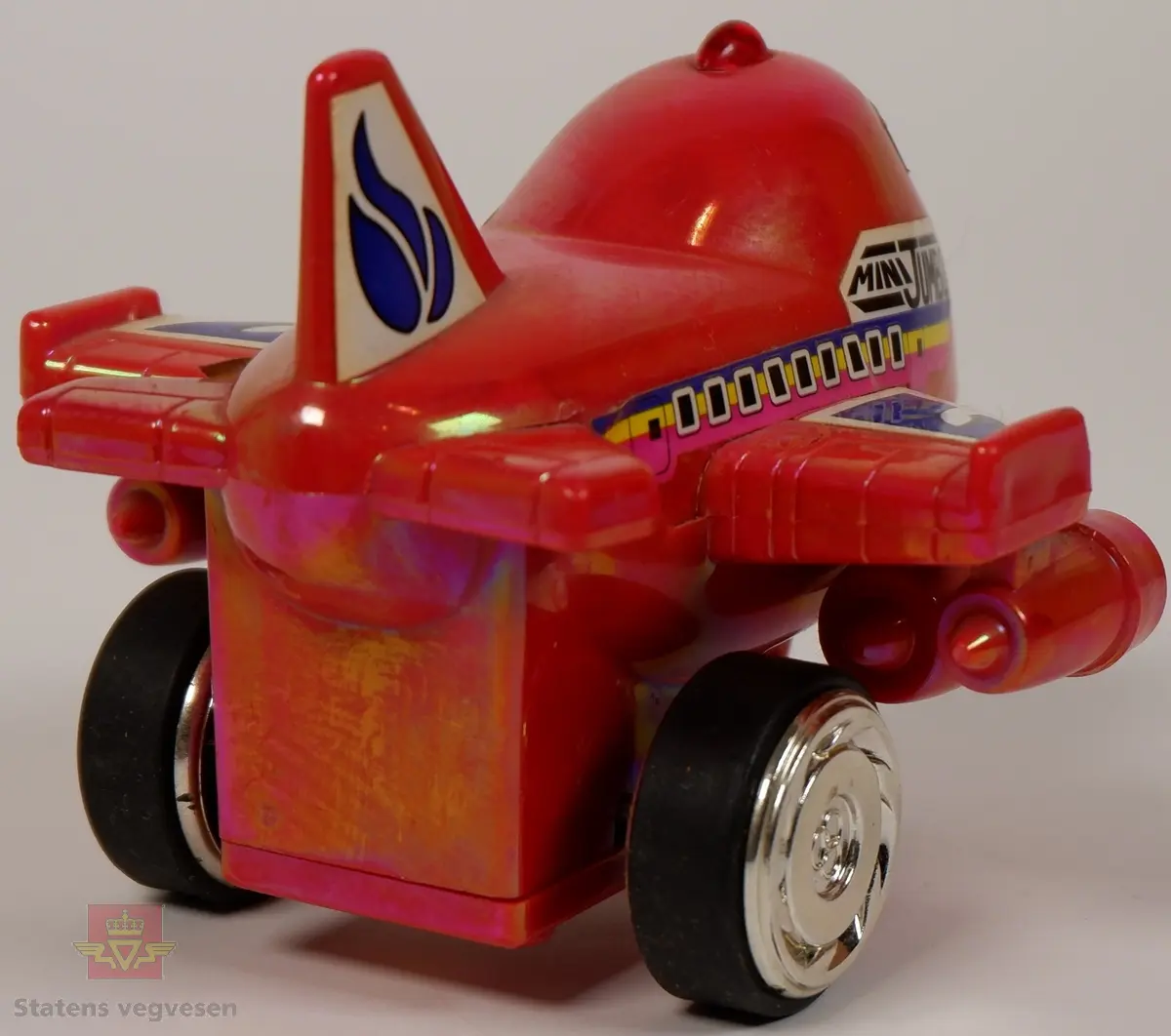 Miniatyrfly i fargen rød med klistremerker som har påskriften MINI JUMBO. Den er utstyrt med en mekaniske som gir den fremdrift når man trekker hjulene bakover.