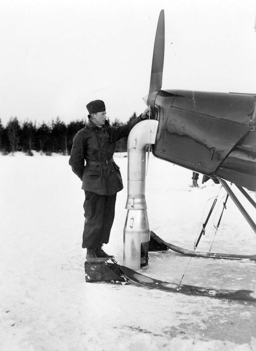 Motorn på flygplan Sk 11 nr 63 värms genom eldning, vintertid. En mekaniker står vid flygplanets nos. Cirka 1932-1939.
