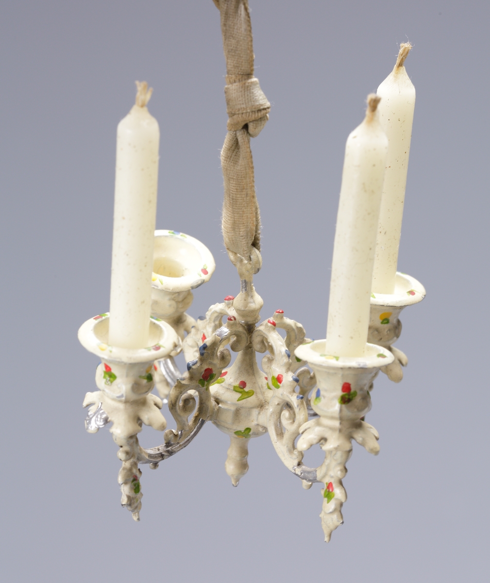 En fyrarmad ljuskrona (för dockskåp) med tre tillhörande vaxljus. Ljuskronan är dekormålad med små blommor och har ett sidenband för upphängning.