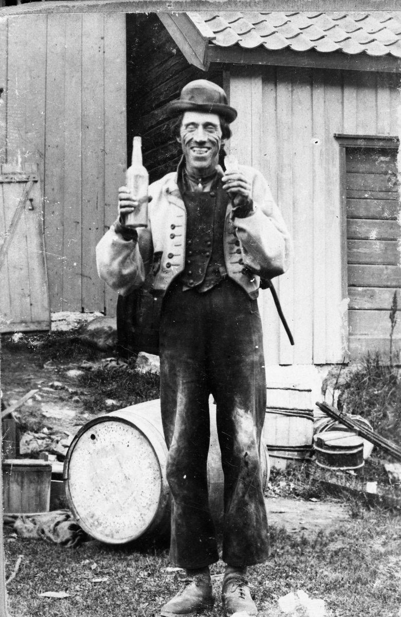 Fotosamling etter Øystein O. Kaasa. (1877-1923). Portrett opprinnelig navngitt, "Brændevinsdjevelen, den søte kløe" av mann i folkedrakt fra Øst Telemark, med flaske.