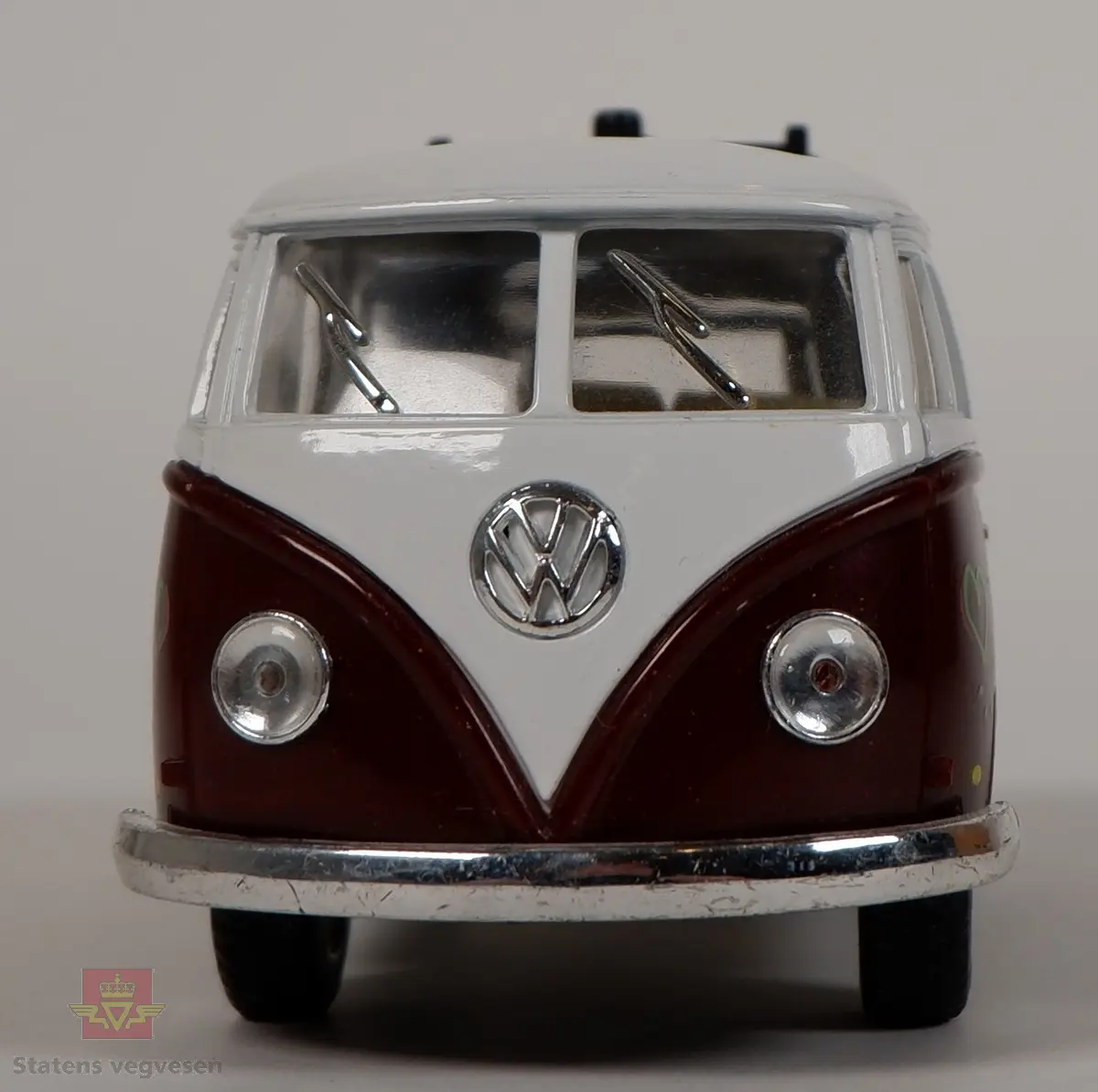 Miniatyrmodell av Volkswagen Transporter T1. Fargen på bilen er hvit og brun, og den er dekorert med hjerter, blomster, fugler og fredstegn i flere farger. Bilen er laget hovedsakelig i metall med plastunderstell og detaljer. Skala 1:32.