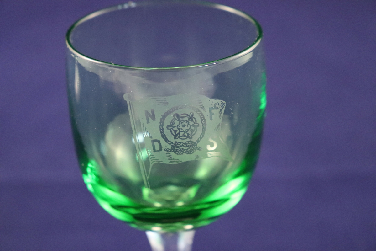 Hetvinsglass med sekskantet stett og rund plate nederst. Glasset er farget grønt med blank stett. Rederiflagget til Det Nordenfjeldske Dampskibsselskab er gravert inn på siden.