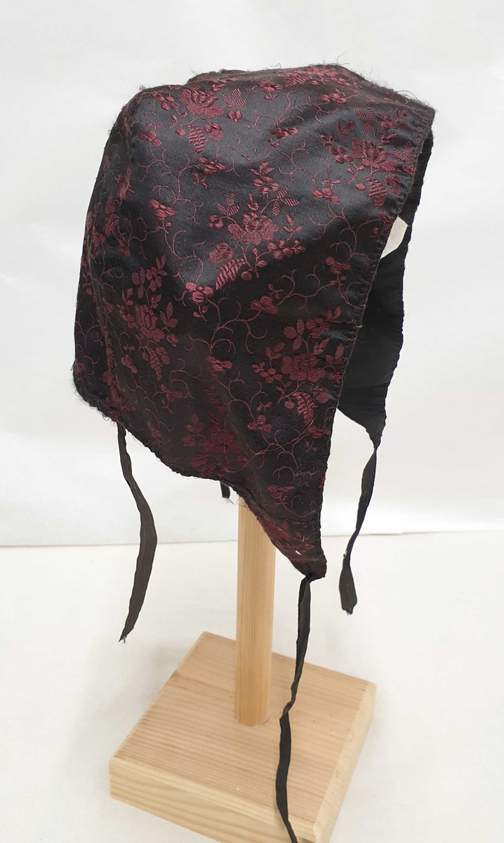 Sydd av svart, blomstret silkedamask med rødfiolette blomster. Fóret med svart linstoff, ripsaktige ca. 1,3 cm brede knytebånd Dobbel rad stikninger i kanten, glatt bakstykke.