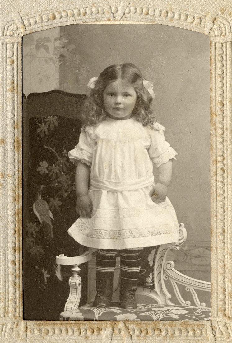 En liten flicka i ljus klänning med spetsinfällningar och mörka, tvärrandiga strumpor. Hon står på en stol med en skärm i bakgrunden. 
Helfigur.