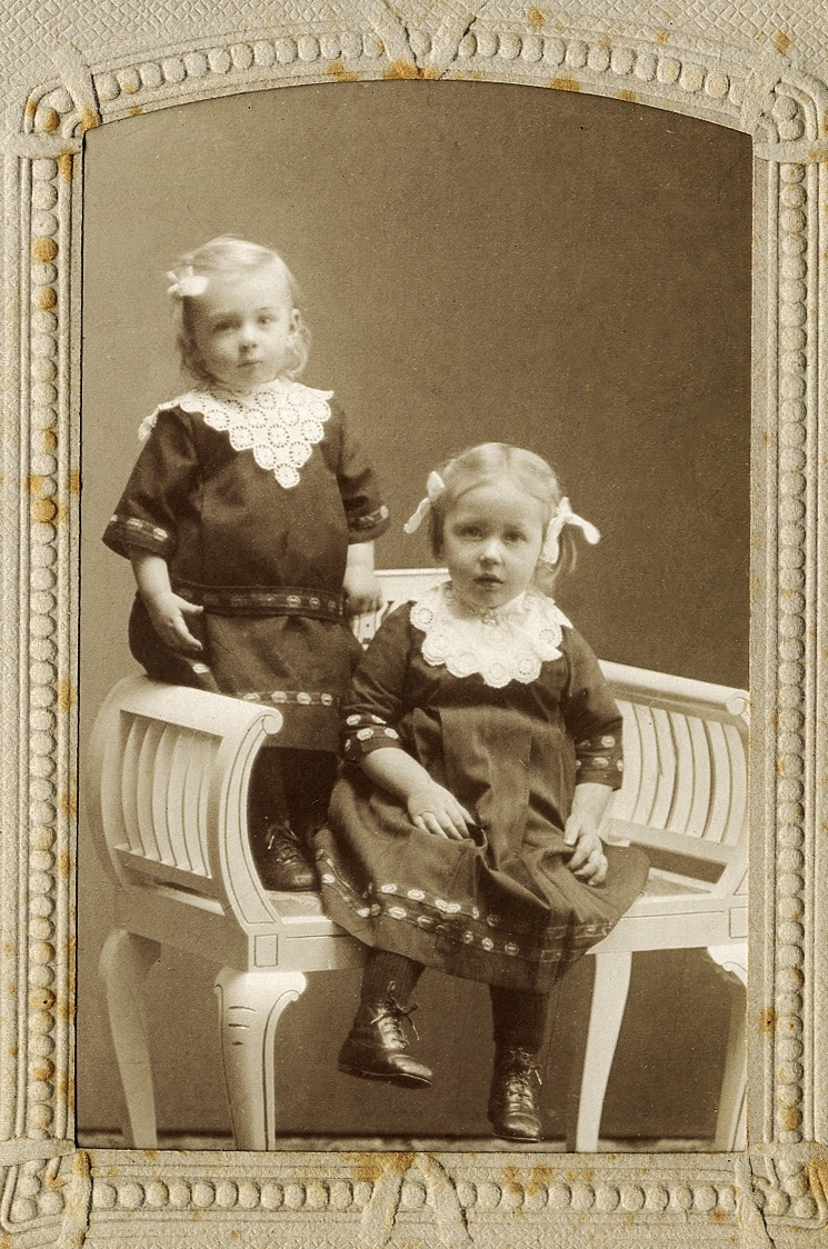 Två små flickor i mörka klänningar med vita spetskragar. 
Helfigur.