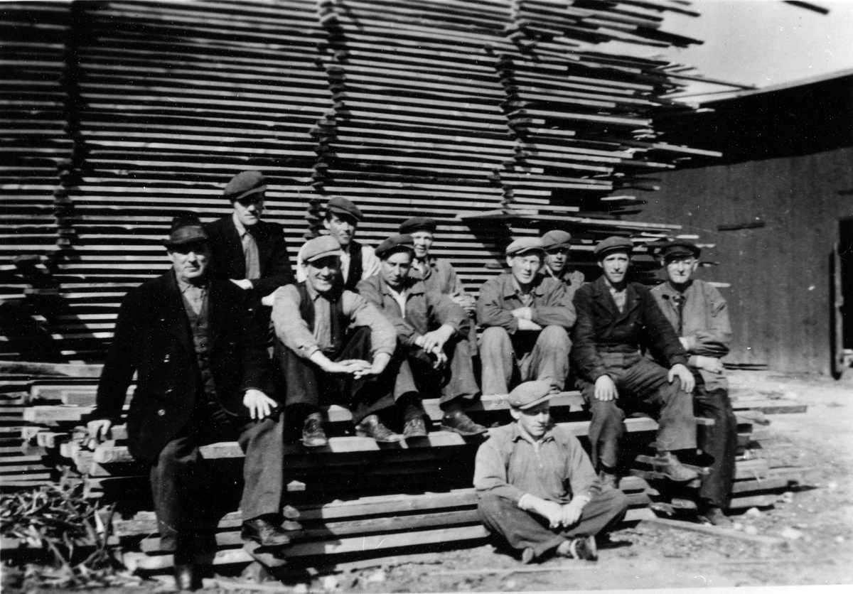 Gruppbild på 11 män som sitter på en hög med bräder framför flera rader av staplade bräder. Alingsås snickerifabrik AB, kv Gjutaren.