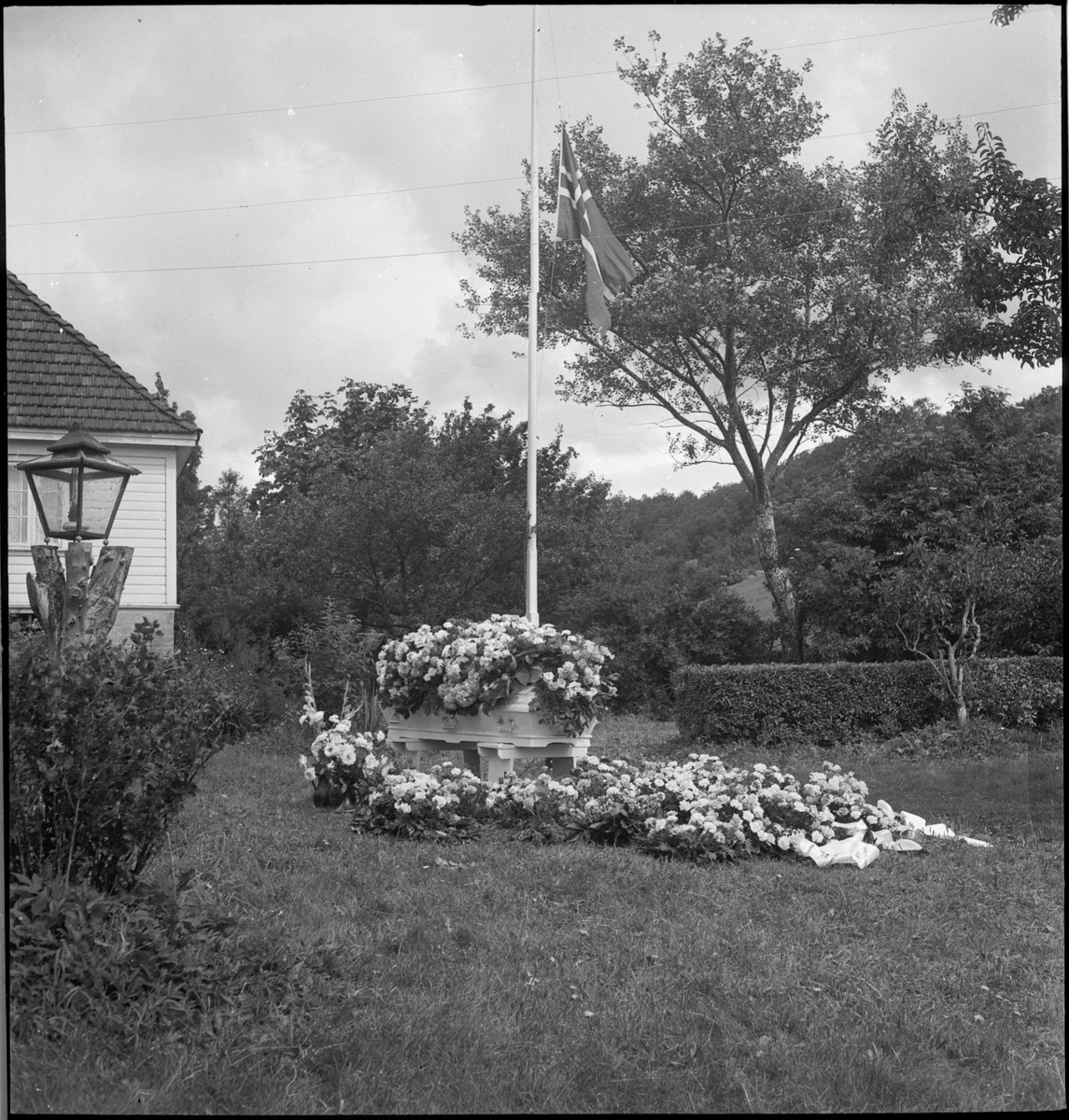 En blomsterkledd kiste i hagen til et bolighus. Flagget er på halv stang og en eldre mann og to gutter er kledd i pentøy i anledning begravelsen.