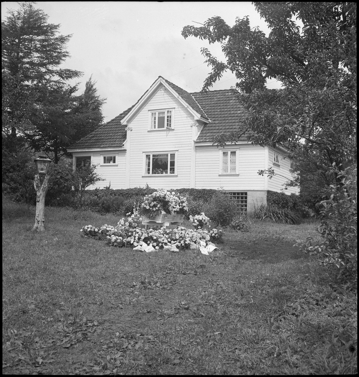 En blomsterkledd kiste i hagen til et bolighus. Flagget er på halv stang og en eldre mann og to gutter er kledd i pentøy i anledning begravelsen.