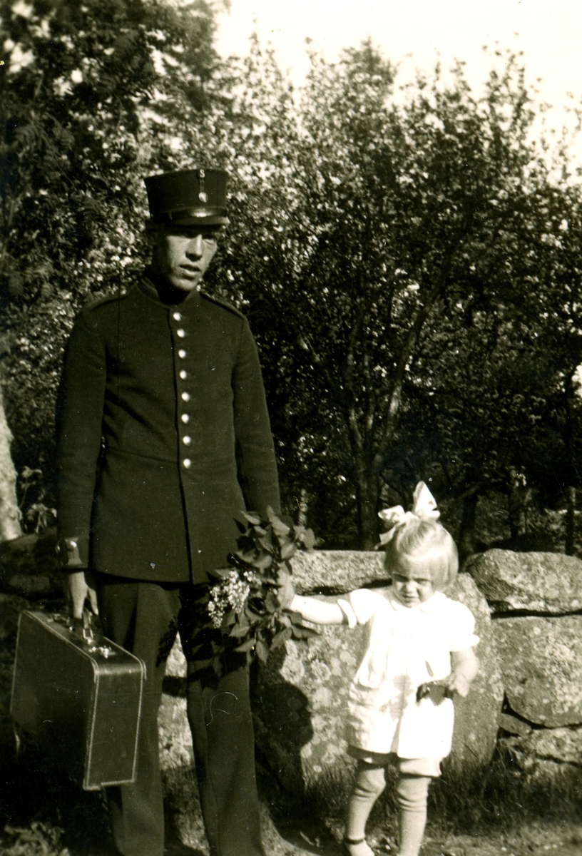 Sporred 1:8 "Fredriks", okänt årtal. En uniformsklädd man står med en resväska i ena handen och en liten flicka i den andra. Flickan har rosett i håret och håller blommor i handen. De står framför en stengärdesgård.