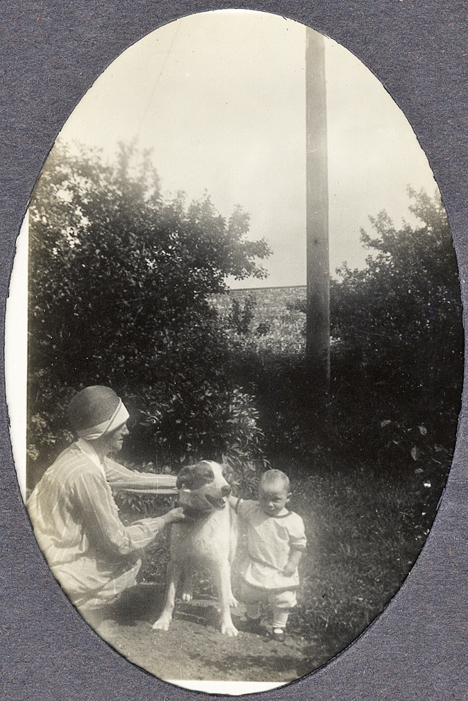 En kvinna i hatt och klänning poserar med en hund och en liten pojke i en trädgård.
