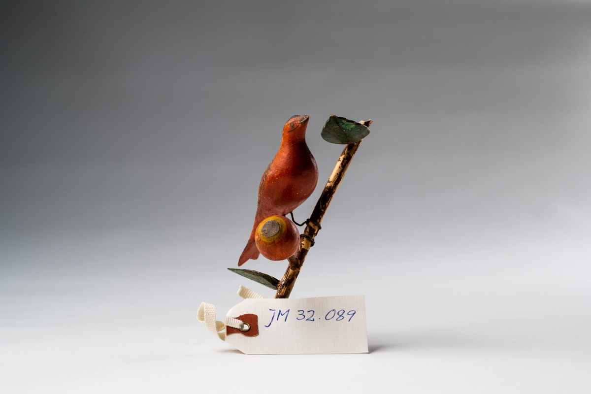 Fågel, leksak (prydnadsföremål), av trä, snidad och bemålad. Den röda fågeln sitter på en sned, brun trädgren med gröna blad och röd-gul frukt (äpple). Alltsammans står på målad bottenplatta i grönt, med dekorativa slingor i vitt, rött, gult och blått. Under bottenplattan är skrivet med blyerts: "1906".