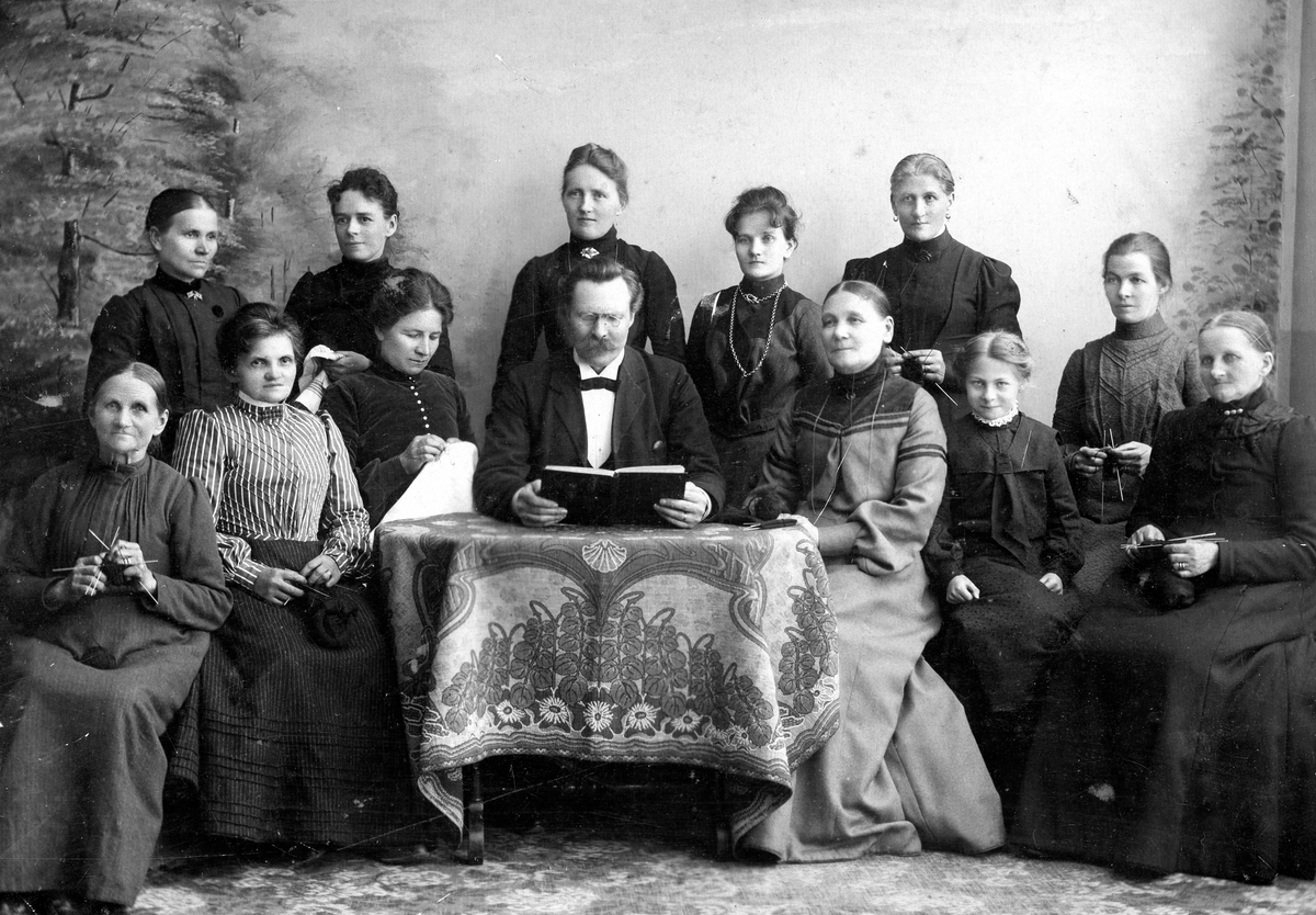 Ateljéporträtt av tolv kvinnor och en man som sitter/står bakom ett litet bord cirka 1900 - 1905. De är från Kållered Missionsförsamlings syförening. Övre raden, andra kvinnan från höger är Maria Svensson från Vommedal Östergård. Övriga elva kvinnor är okända. I mitten sitter mannen vid bordet och håller en öppen bok i sina händer.