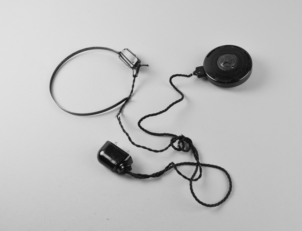 Høreapparat i bakelitt bestående av en rund mikrofon med klips bak til feste, en hodebøyle med en liten høytaler som plasseres utenfor det ene øret. En tvunnet snorledning forbinder mikrofon og høytaler med en stikkontakt som skal tilsluttes et batteri (batteri mangler).