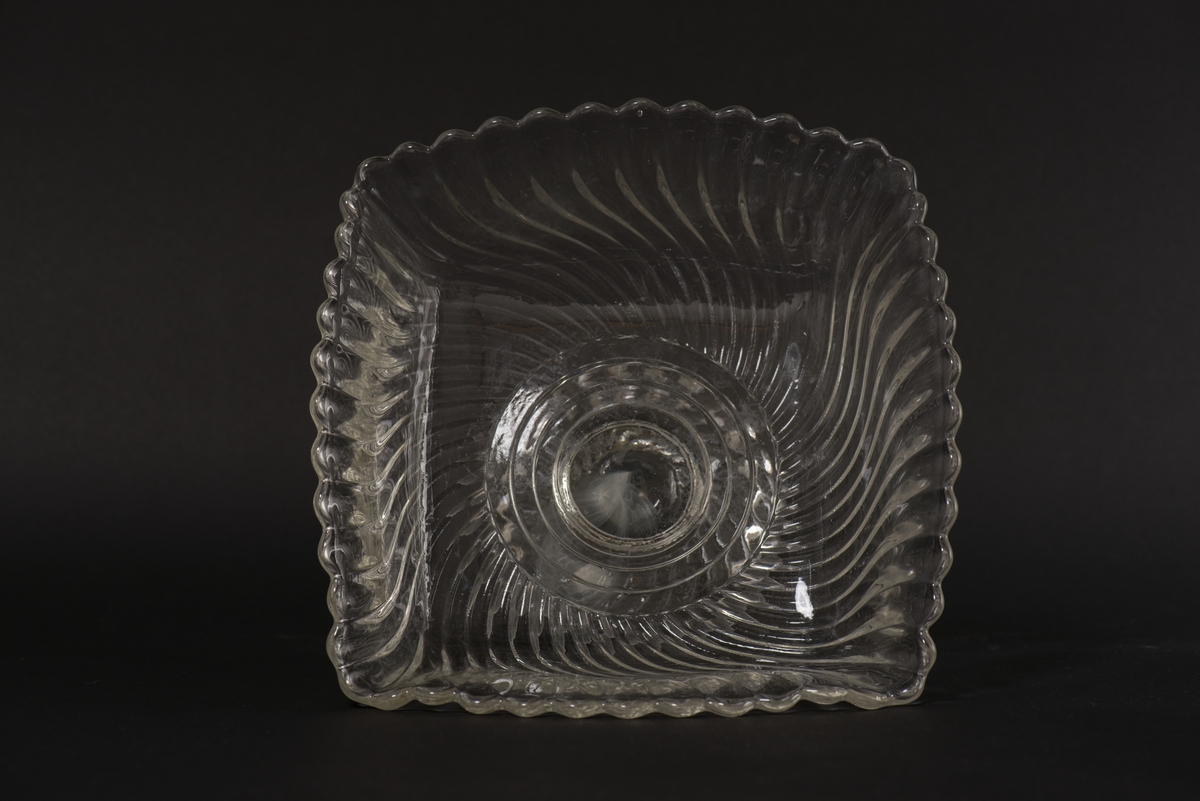 Fyrsidig skål på fot av ofärgat pressglas. Pressad yttäckande dekor av spiralvridna åsar som avslutas i en vågformad kant.