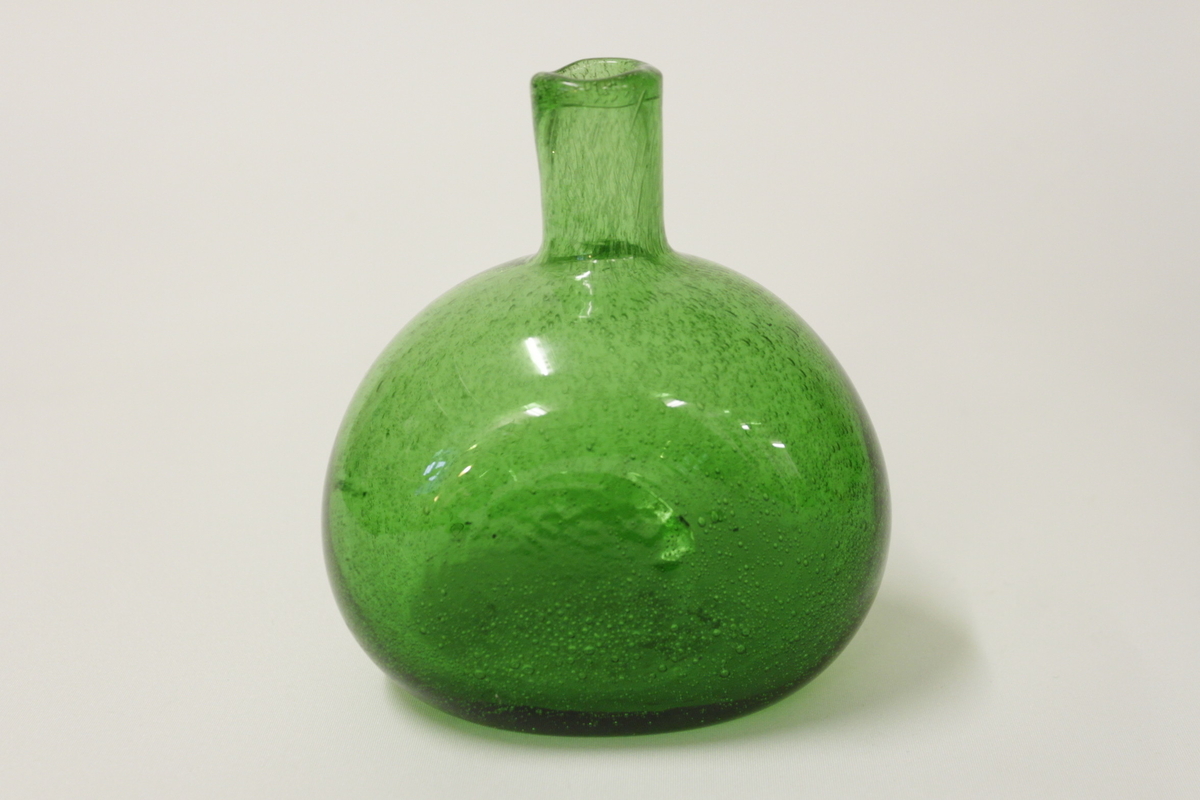 Glasflaska använt i bikupa för socker och vatten. Bullig grön glasflaska med ena sidan slät.