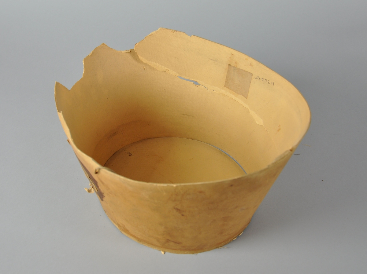 Oval hatteekse av papp. med lokk. Del av esken er revet av på den ene siden. Lokket er dekorert med mynter og laurbærblader.