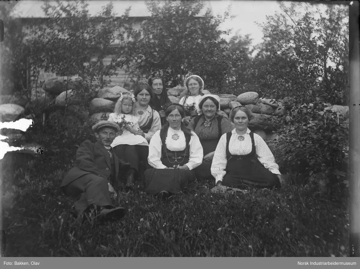 Gruppe mennesker sittende på gresset foran steinmur. Laftet bygg i ...