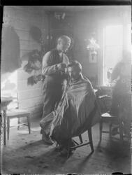 Mann sittende på stol i bolig og blir klippet av frisør. Man