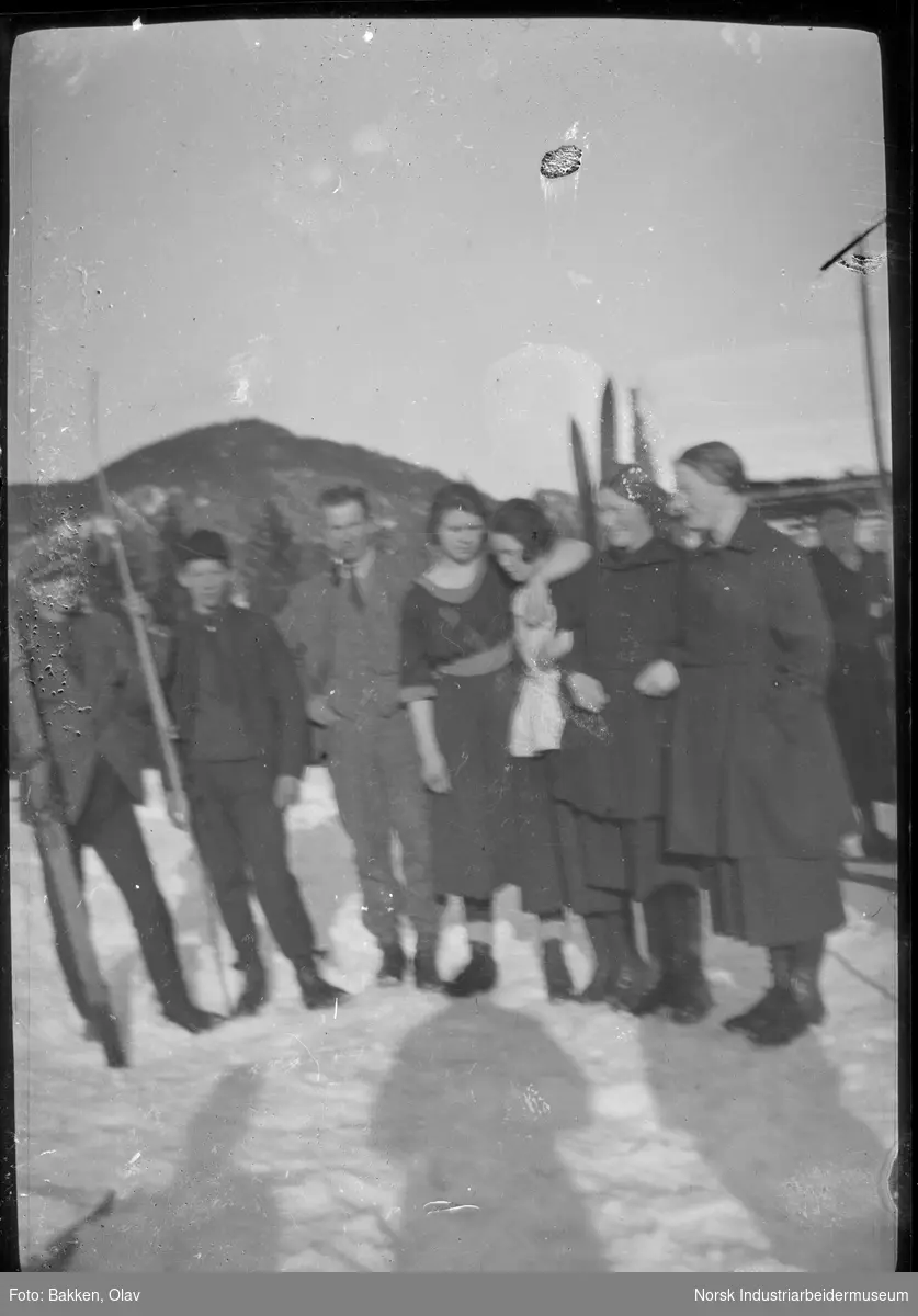 Voksne og ungdom stående oppstilt utendørs i snø, fler med ski i hånden.