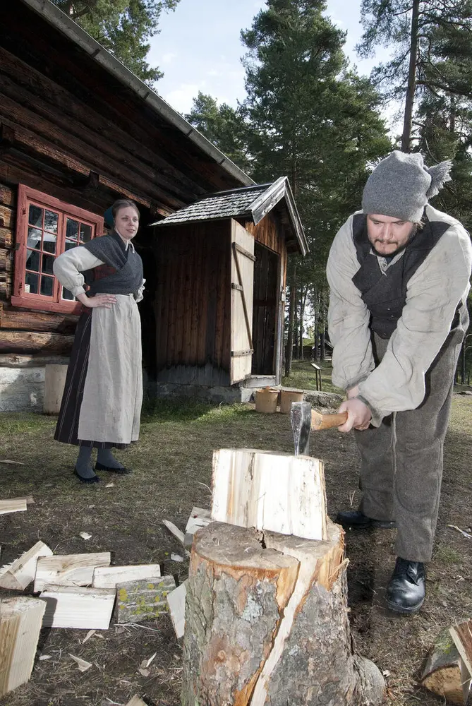 En husmann med grå klær står utenfor husmannsplassen sin og hogger ved. Husmannskona med stakk, skaut, forkle og omslagssjal står med hendene i sida og ser på.