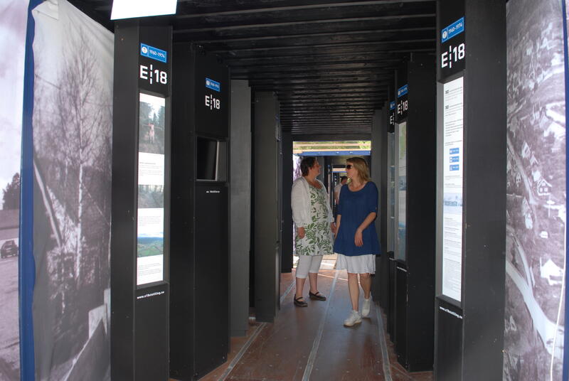 Plansjer med tekst og bilde om E18 inne i en container. To personer ser på utstillingen.